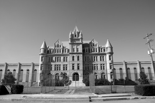 Historic stone building with turrets, surrounded by a fence, under a clear sky in black and white.