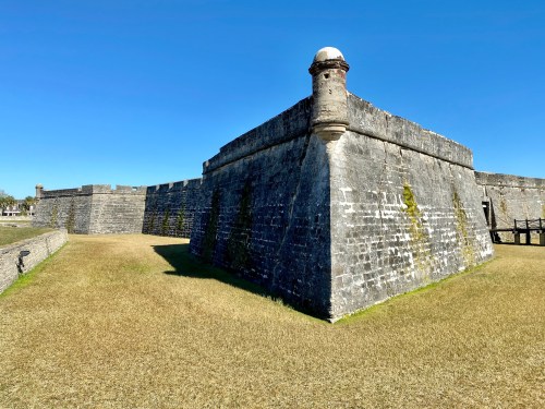 A historic stone fort with a rounded tower, set against a clear blue sky and grassy landscape.