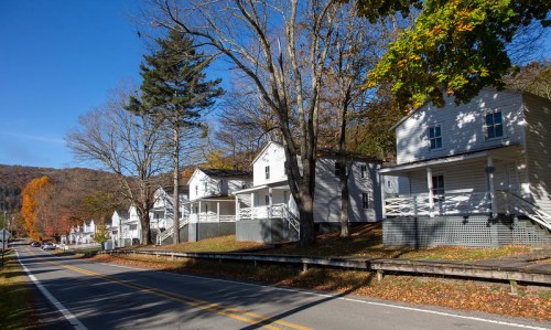 A row of white houses with porches along a tree-lined road, surrounded by autumn foliage and mountains in the background.
