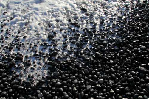 A close-up of smooth black pebbles on a beach, with white foam from waves washing over them.