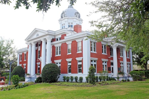 Historic red brick building with white columns and a dome, surrounded by greenery and a well-maintained lawn.
