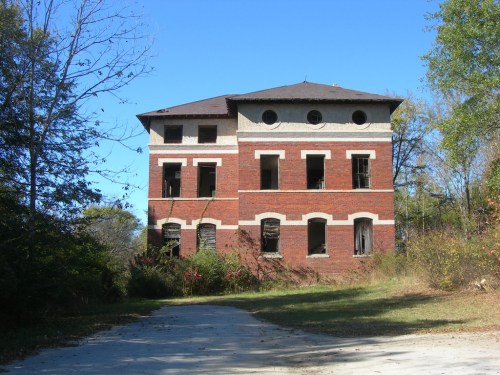 Abandoned brick building with broken windows, surrounded by overgrown vegetation and a clear blue sky.