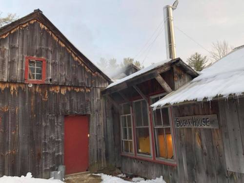 A rustic sugar house with a red door and smoke rising from a chimney, surrounded by snow and trees.