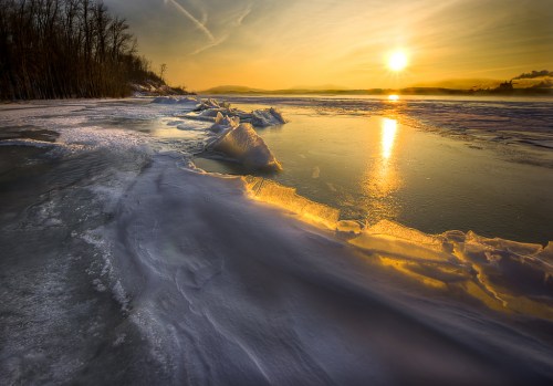 A serene winter landscape at sunset, featuring icy shores and a golden reflection on the water.