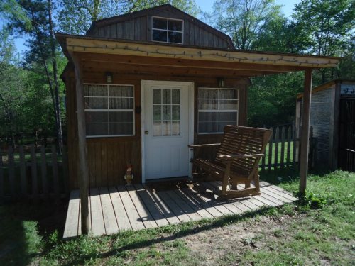 A small wooden cabin with a porch and rocking chair, surrounded by greenery and a wooden fence.