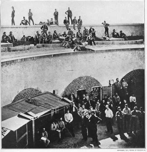 Historic black-and-white photo of a large group of men gathered in a courtyard, some seated on walls and others standing.