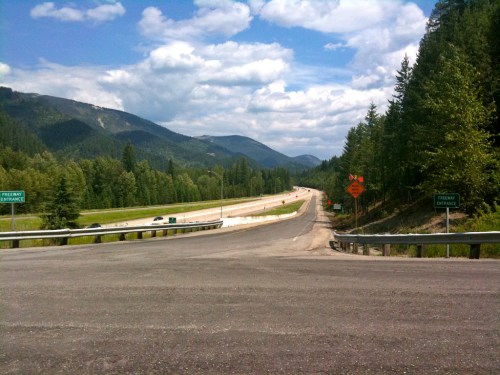 A wide highway stretches into the distance, flanked by green trees and mountains under a partly cloudy sky.