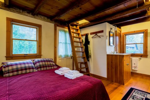 Cozy bedroom with a red quilt, wooden beams, a ladder, and a small bathroom area with towels neatly arranged.