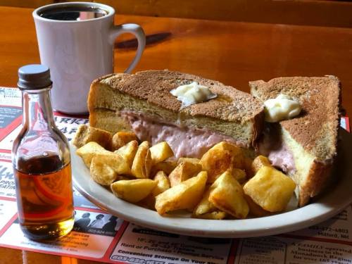 A plate with a sandwich filled with pink spread, served with golden potato wedges and a cup of coffee.