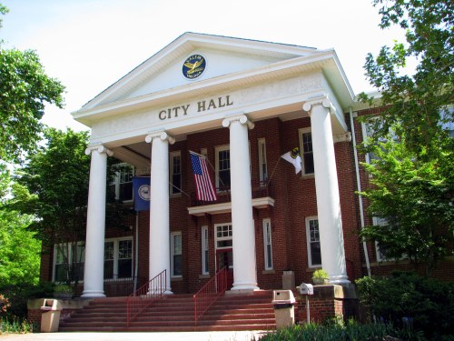 A brick city hall building with white columns, flags, and greenery surrounding it on a sunny day.