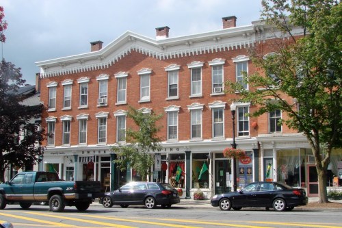 Historic brick building with multiple windows, shops on the ground floor, and parked cars in front.