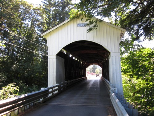 A white covered bridge surrounded by trees, leading into a shaded pathway.