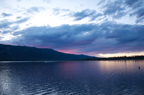 A serene lake at sunset, reflecting purple and blue hues in the sky, with mountains in the background and calm water.