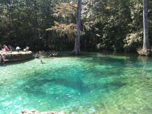 A clear, turquoise spring surrounded by trees, with people swimming and relaxing on the shore.