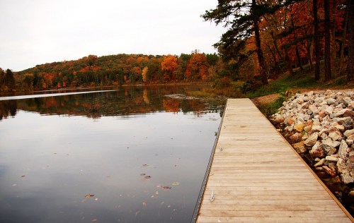 A wooden dock extends over a calm lake, surrounded by trees displaying vibrant autumn colors.