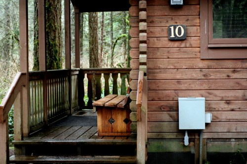 A wooden cabin with a porch, featuring a bench and the number 10 on the wall, surrounded by trees.