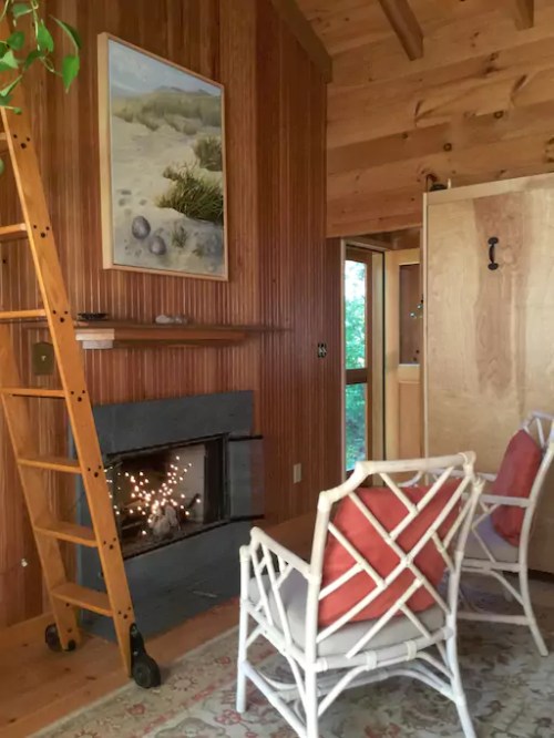 Cozy interior with wooden walls, a fireplace, a ladder, and two white chairs with red cushions. A painting hangs above the mantel.