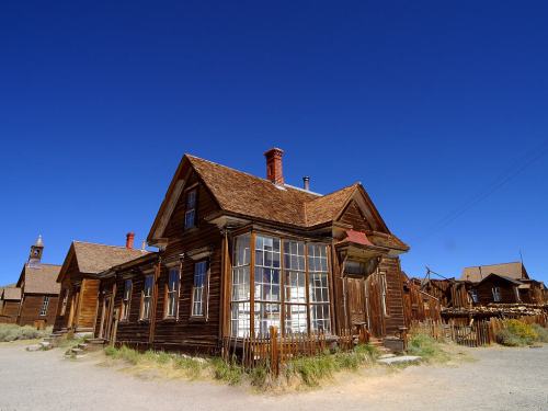 A historic wooden building with a sloped roof, surrounded by a dusty landscape under a clear blue sky.