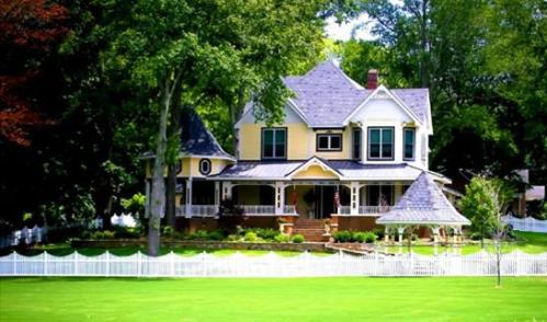 A charming yellow Victorian house with a large porch, surrounded by trees and a white picket fence on a green lawn.