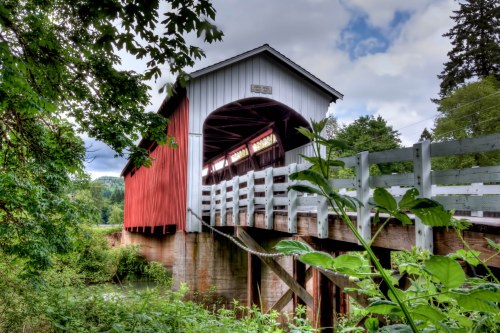 A red and white covered bridge spans a river, surrounded by lush greenery and a cloudy sky.