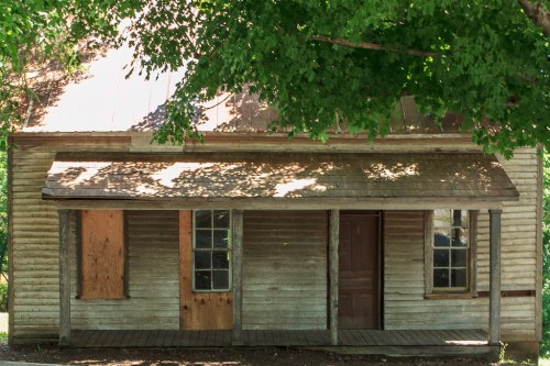 A weathered wooden house with boarded windows, a porch, and a tree casting shade in front.