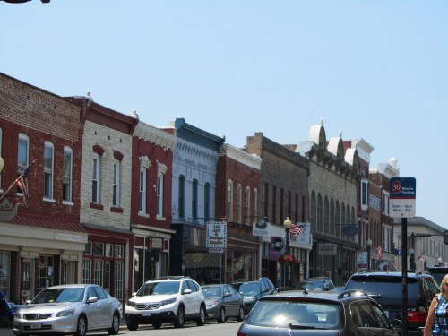 A charming street lined with historic buildings and parked cars, featuring colorful storefronts and American flags.