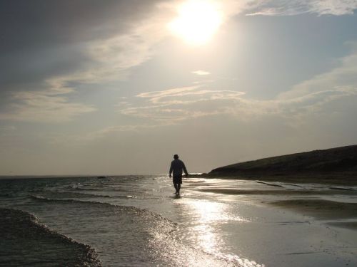 A person walks along a beach at sunset, with shimmering water and a cloudy sky in the background.