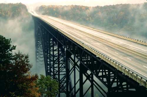A high bridge spans a foggy valley, surrounded by trees with autumn foliage. The road is empty and wet from rain.