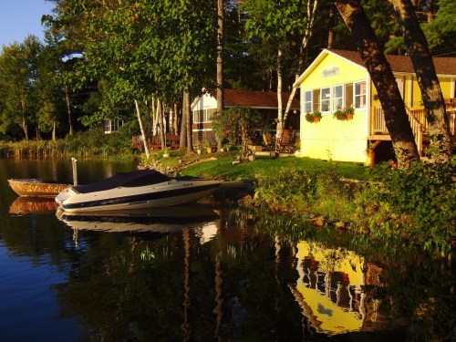 A serene lakeside scene featuring a yellow cottage, a boat, and trees reflecting in the calm water.