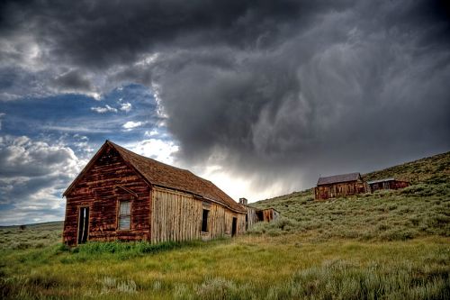 Abandoned wooden buildings on a grassy hillside under a dramatic, cloudy sky.