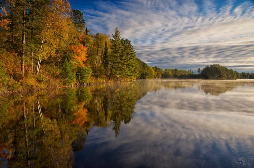 A serene lake reflects colorful autumn trees and a cloudy sky, creating a peaceful natural landscape.