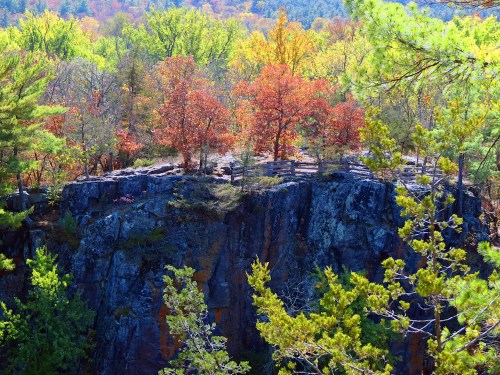 A rocky cliff surrounded by vibrant autumn trees in shades of red, orange, and yellow under a clear blue sky.