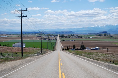 A long, straight road stretches into the distance, flanked by fields and mountains under a partly cloudy sky.