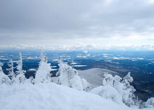 A snowy mountain landscape under a cloudy sky, with distant hills and valleys blanketed in white.