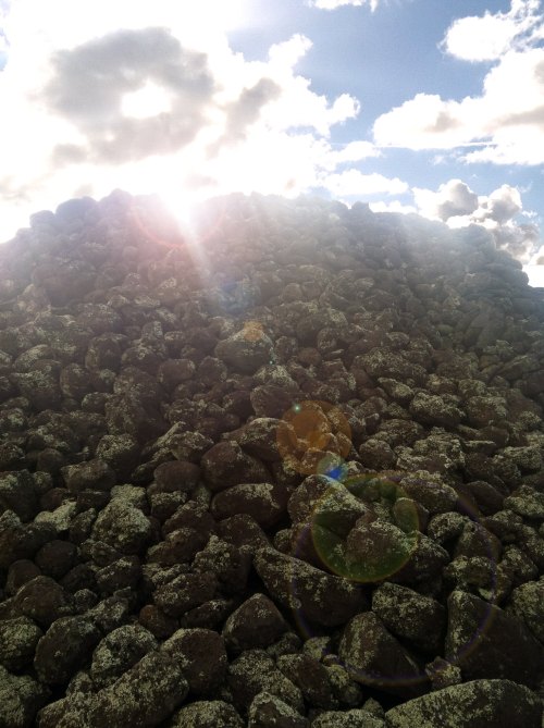 A pile of dark rocks under a bright sky with clouds, sunlight creating a lens flare effect.