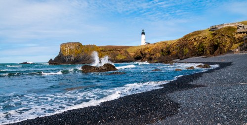 A scenic view of a lighthouse on a rocky coastline, with waves crashing on a black pebble beach under a blue sky.