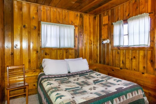 Cozy wooden cabin bedroom with a double bed, quilted blanket, and a chair by the window. Natural light filters in.