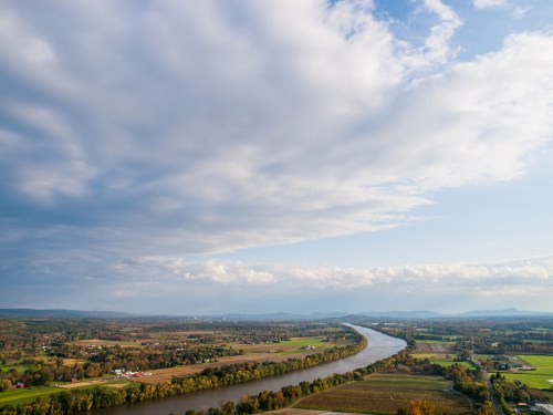 A scenic view of a winding river surrounded by lush fields and distant mountains under a partly cloudy sky.