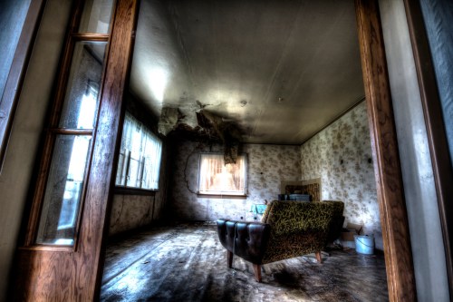 An abandoned room with a worn sofa, peeling wallpaper, and a ceiling showing signs of damage and decay.