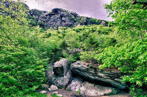 Lush green trees and large rocks at the base of a steep, rocky cliff under a cloudy sky.