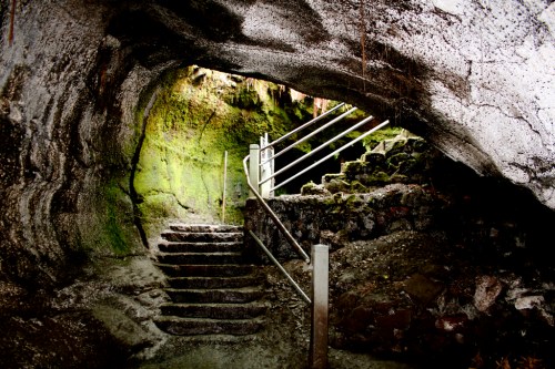 A moss-covered cave entrance with stone steps and metal railing leading into a dimly lit interior.