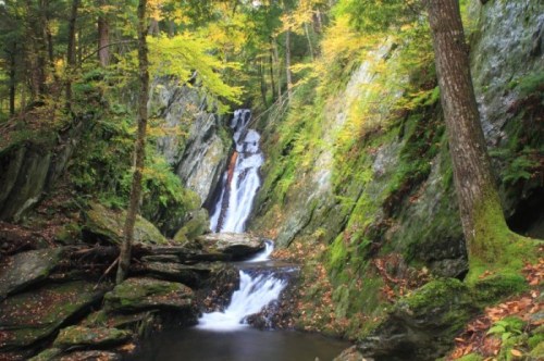 A serene waterfall cascades down rocky cliffs, surrounded by vibrant autumn foliage and lush greenery.