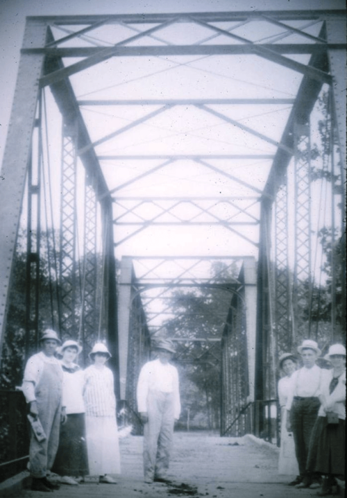 A group of people in vintage clothing stands on a bridge, surrounded by trees, in a black-and-white photo.