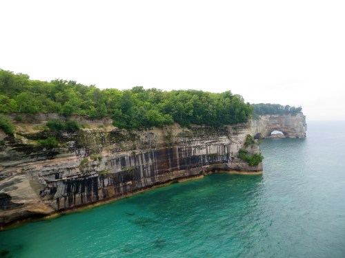 A rocky cliff with lush greenery overlooks clear turquoise waters and a natural arch in the distance.