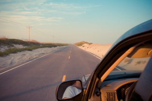 A view from a car window of an empty road lined with sand dunes under a clear sky.