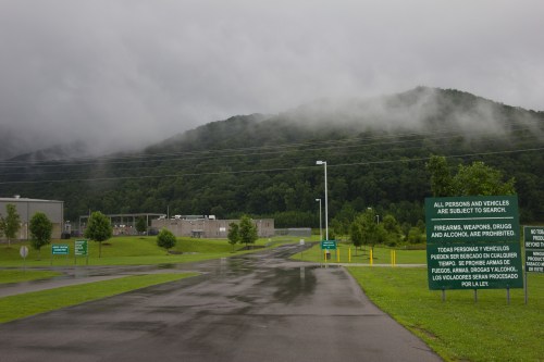 A cloudy landscape with a road leading to a facility, surrounded by green hills and warning signs about searches.