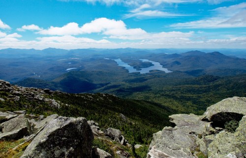 A panoramic view of mountains and a lake surrounded by lush greenery under a blue sky with scattered clouds.