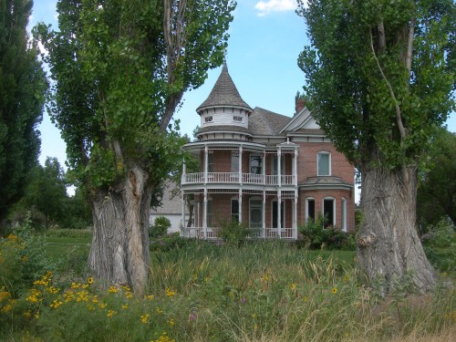 A historic Victorian-style house surrounded by tall trees and colorful flowers in a lush garden setting.