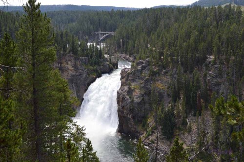 A scenic view of a waterfall cascading through rocky cliffs, surrounded by lush green trees and a distant bridge.