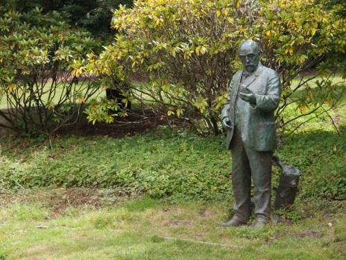 A bronze statue of a man in a suit holding a cane, standing in a grassy area surrounded by bushes.